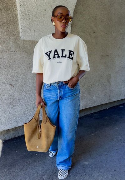 Camiseta gráfica blanca con "YALE" en negro, jeans de mezclilla azul, bolso de cuero marrón claro y zapatos de lona a cuadros, colocados frente a una pared texturizada.