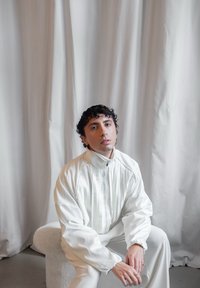 Weiße Jacke mit vertikalen Streifen, lässiger Schnitt, hoher Kragen und elastischen Bündchen. Die Person sitzt auf einem hellen, strukturierten Stuhl vor einem drapierten Hintergrund.