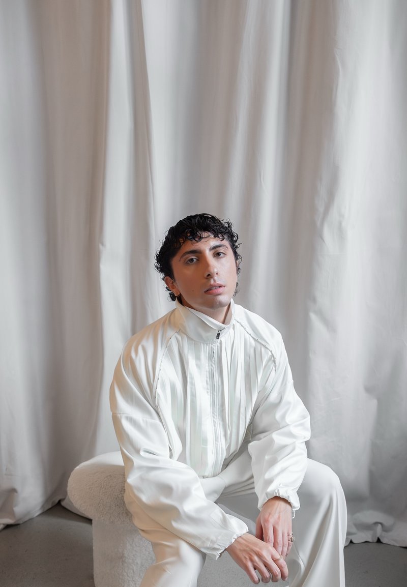 Jacket branco com riscas verticais, corte descontraído, gola alta e punhos elásticos. A pessoa está sentada numa cadeira de textura leve contra um fundo drapeado.