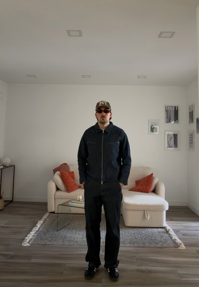 Hombre con chaqueta oscura, pantalones, gafas de sol y gorra, de pie en una sala de estar moderna con sofá crema, mesa de cristal y cuadros enmarcados en la pared.