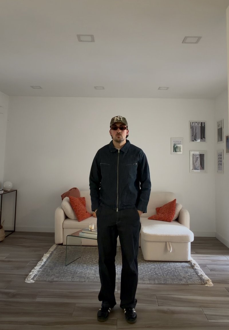 Mand iført mørk jakke, bukser, solbriller og kasket står i en moderne stue med cremefarvet sofa, glasbord og indrammede billeder på væggen.