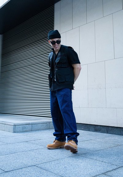 Chaleco negro de múltiples bolsillos, camisa negra de manga corta, jeans azules de corte holgado, botas de trabajo marrones, de pie sobre una superficie de piedra con una pared gris detrás.