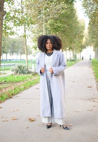 Γκρι μακρύ παλτό με μεγάλα πέτα, φορεμένο πάνω από γκρι τοπ και παν trousers φαρδιάς γραμμής. Μαύρες ψηλοτάκουνες γόβες. Σγουρά μαλλιά, εξωτερικός χώρος.