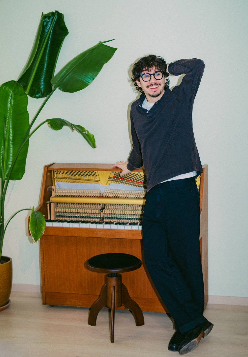 Eiket oppreist piano med synlige mekaniske deler, ledsaget av en svart krakk. En stor grønn plante står i bakgrunnen mot en lys vegg.