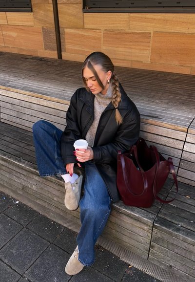 Chaqueta de cuero negra, suéter gris, jeans de pierna ancha, zuecos beige, calcetines blancos, sosteniendo una taza de café blanca, bolsa de mano burdeos, banco de madera.