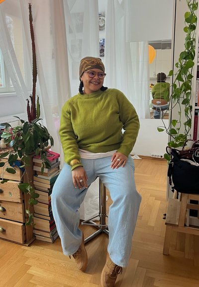 Persona con gafas, suéter verde y gorro marrón sentada en un taburete en una habitación luminosa con plantas y libros cerca.