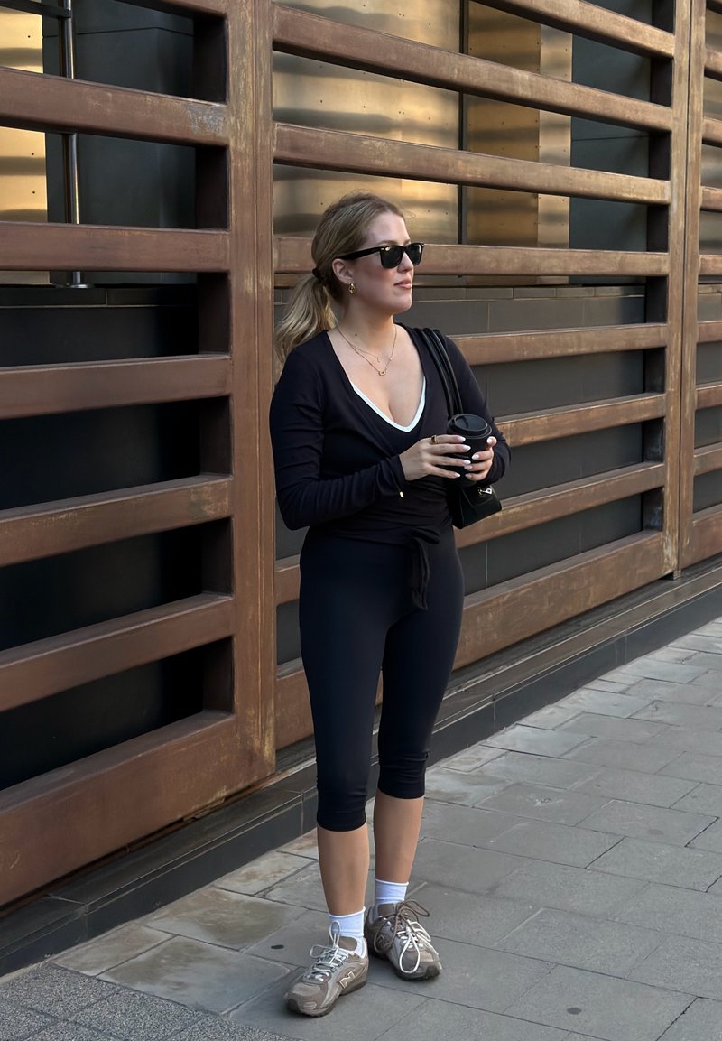 Femme portant une tenue de sport noire et des lunettes de soleil, debout sur le trottoir, tenant un gobelet de café à emporter près d’un mur en bois à lattes.
