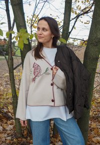 Beež kardigan, millel on tikitud linnu muster, burgundia nupud, selga pandud valge särgi ja pruuni vatitud jope peale, mis on kombineeritud siniste teksadega.