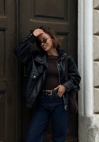 Zwarte leren jacket met grote revers, bruin getailleerd topje, hooggekeurde donkerblauwe jeans en een gestructureerde bruine handtas. Ronde zonnebril.