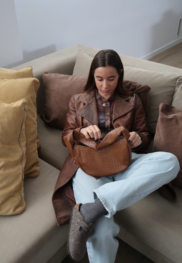 Brown woven leather bag with a slouchy design, placed in hands of a person wearing a brown leather coat, light blue jeans, and grey socks.