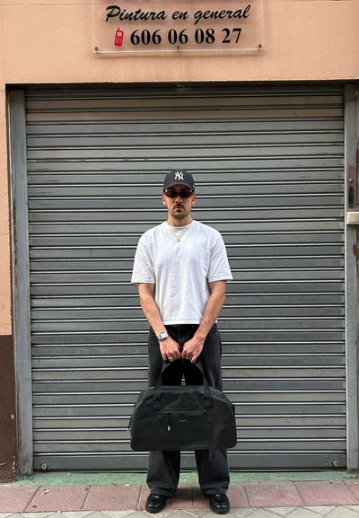 Bolsa de deporte sintética negra con forma redondeada y asas dobles en la parte superior. El modelo lleva una camiseta blanca y pantalones negros, de pie frente a una persiana gris.