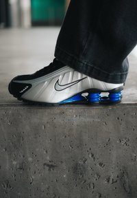Schwarze und silberne Nike-Sneaker mit blauem gepolstertem Sohlen, die eine strukturierte Oberfläche, schwarze Schnürsenkel und ein markantes Logo an der Seite aufweisen.