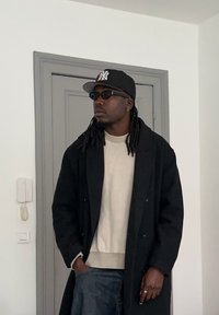 Fekete kétsoros kabát világos bézs pulóver felett, söt�ét farmernadrággal és fekete sapkával. Rasztaszemek, napszemüveg és gyűrű teszi teljessé a megjelenést.