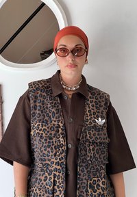 Ženska nosi adidasov telovnik s leopardjim vzorcem čez rjavo srajco, oranžno rutico, sončna očala in plastične biserne verižice, stoji ob beli steni.
