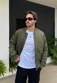 Maslinasto zelena bomber jakna s glatkom teksturom, rebrastim manžetama, prednjim džepom, složena preko bijele majice, u kombinaciji s crnim hlačama.