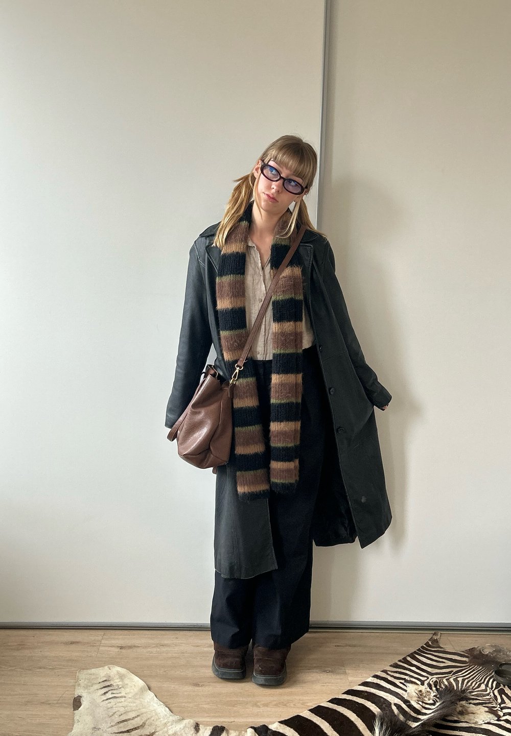 Schwarzer langer Mantel über einem beigen Oberteil, schwarze Hose und braune Stiefel. Ein gestreifter Schal in Beige, Braun und Grün sowie eine braune Umhängetasche.
