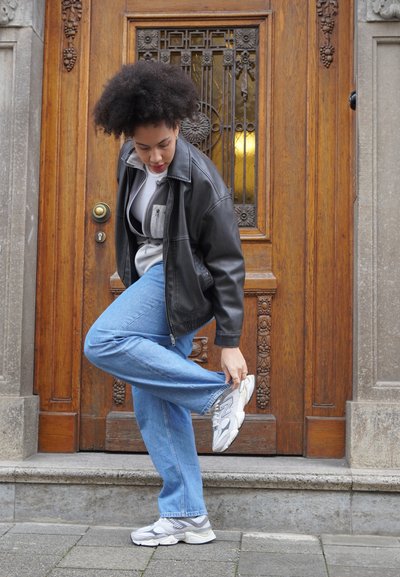 Chaqueta de cuero negra sobre una sudadera gris con cremallera, jeans azules y zapatillas grises en una persona que está de pie junto a una puerta de madera con detalles ornamentales.