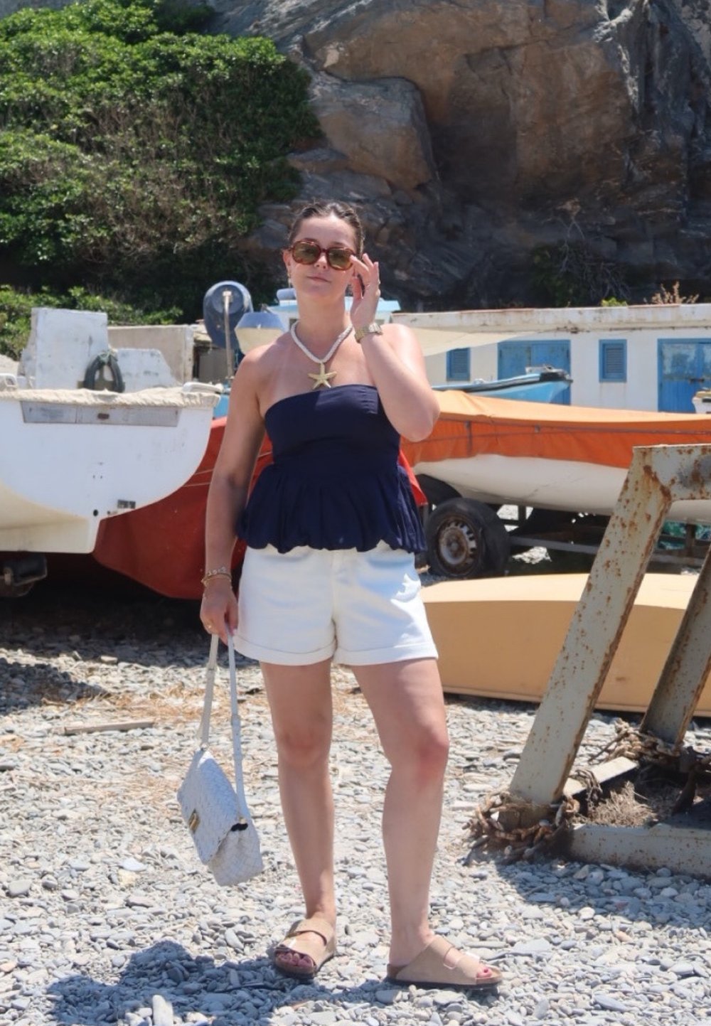 Μπλε peplum τοπ, λευκά σορτς, καφέ σανδάλια και μια ανοιχτόχρωμη τσάντα. Κολιέ με αστερία και γυαλιά ηλίου. Λιθόστρωτο έδαφος, βάρκες στο φόντο.