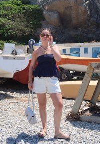 Μπλε peplum τοπ, λευκά σορτς, καφέ σανδάλια και μια ανοιχτόχρωμη τσάντα. Κολιέ με αστερία και γυαλιά ηλίου. Λιθόστρωτο έδαφος, βάρκες στο φόντο.