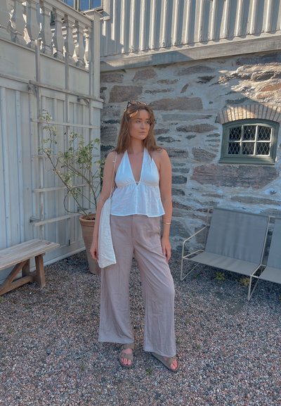 Top halter blanco y pantalones anchos beige con una textura fluida. Pared verde grisáceo, banco de madera y planta en maceta al fondo.
