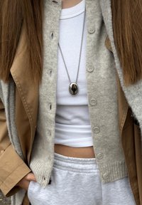 Persoană purtând un palton bej în straturi și un cardigan gri peste un top alb fără mâneci, pantaloni gri de trening și un colier cu pandantiv oval argintiu.
