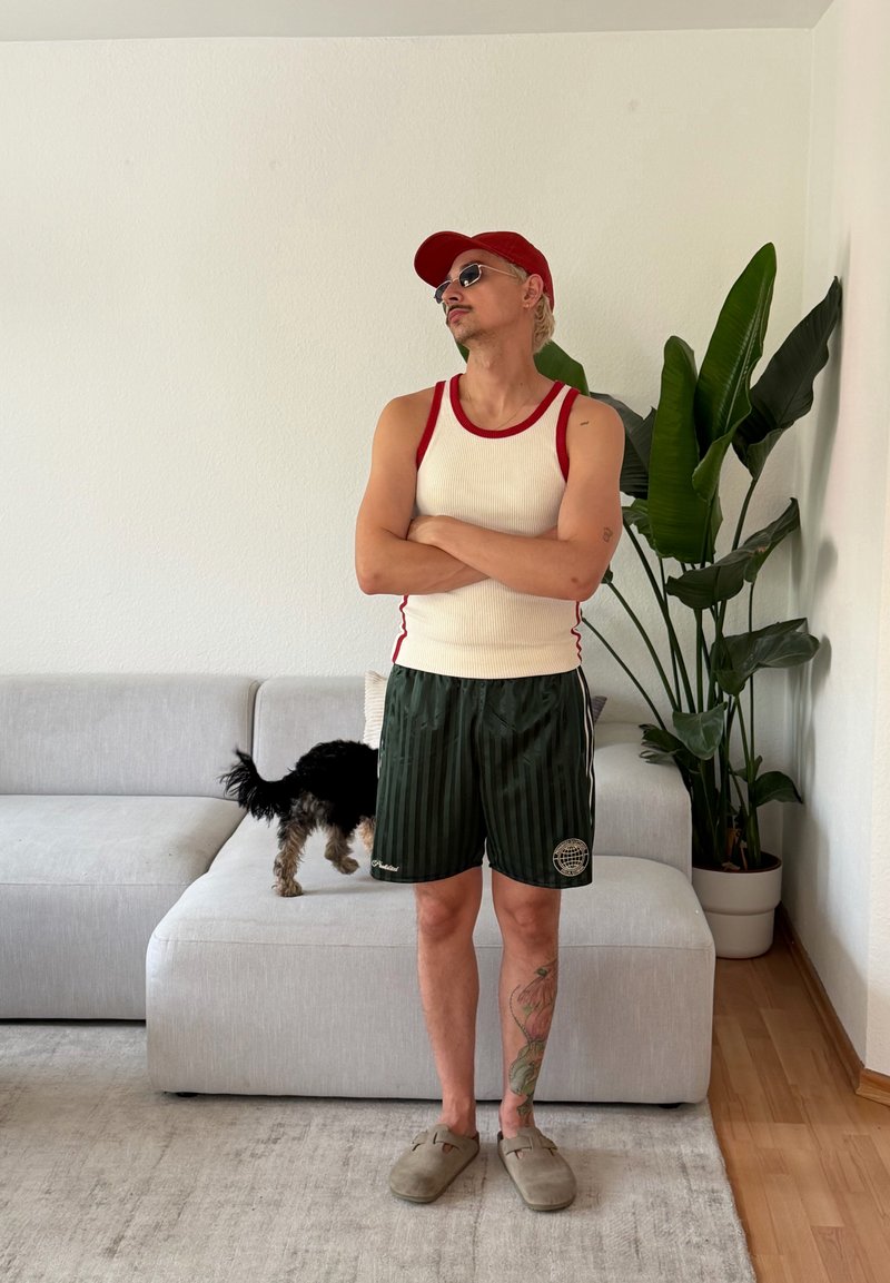 Homme portant des lunettes de soleil, une casquette rouge, un débardeur blanc et un short noir, debout, les bras croisés dans le salon près d’un chien sur le canapé et une grande plante en pot.