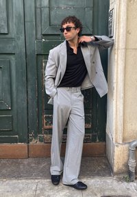 Muž v sivom obleku a čiernej košeli so slnečnými okuliarmi, stojaci pri opotrebovaných zelených dverách s jednou rukou v vrecku a druhou pri krku.