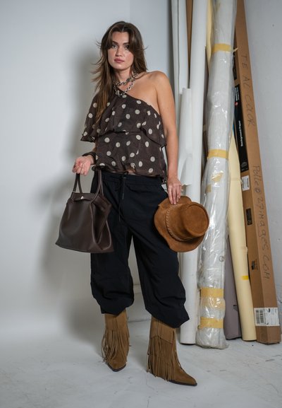 Mujer con un top de un solo hombro con lunares, pantalones negros, botas con flecos, sosteniendo un sombrero marrón y un bolso marrón oscuro, de pie junto a lienzos enrollados.