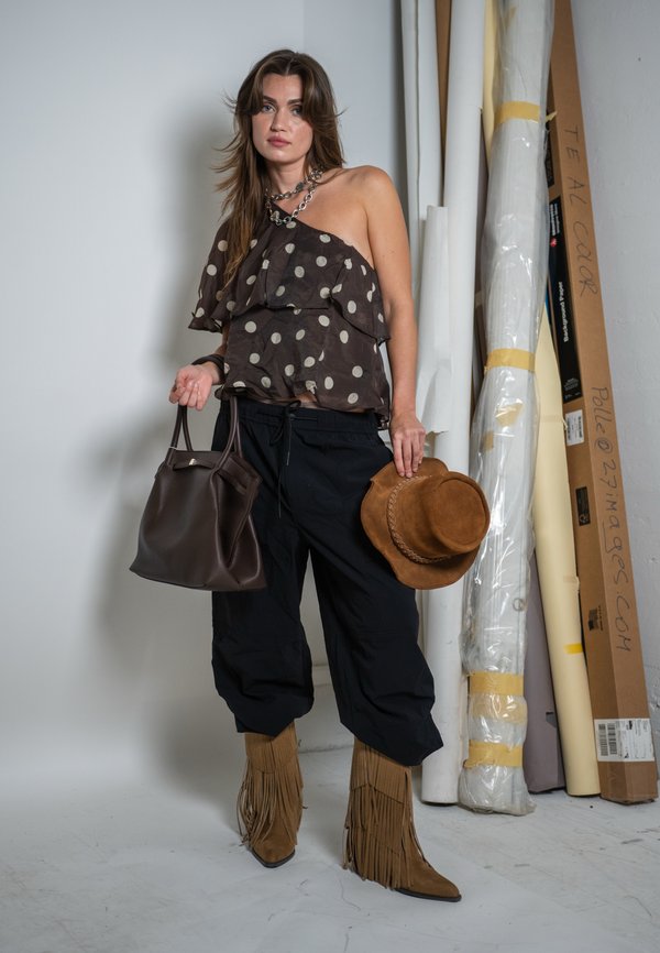 Frau trägt ein gepunktetes One-Shoulder-Oberteil, schwarze Hose, Fransenstiefel, hält einen braunen Hut und eine dunkelbraune Handtasche, steht neben aufgerollten Leinwänden.