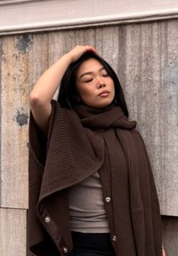 Écharpe en tricot marron avec une texture côtelée, stylisée en châle. Présente un col haut et des accents de boutons décoratifs. Un haut gris visible en dessous.