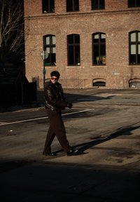 Homme portant une veste en cuir sombre, des gants et des lunettes de soleil, traversant une rue ensoleillée près d'un bâtiment en brique avec des fenêtres cintrées.