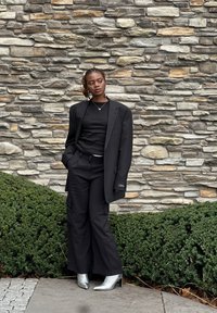Zwarte oversized blazer over een zwart top en zwarte cargobroek, gecombineerd met zilveren enkellaarsjes. Textuurvolle stenen muur op de achtergrond.