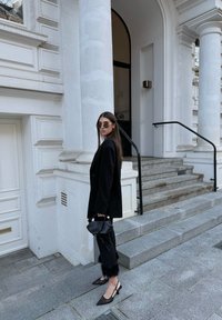 Μαύρη σακάκι πάνω από εφαρμοστό τοπ, ταγιέρ μαύρο παντελόνι με γούνινες λεπτομέρειες, μυτερές μαύρες slingback γόβες και στρογγυλά γυαλιά ηλίου.