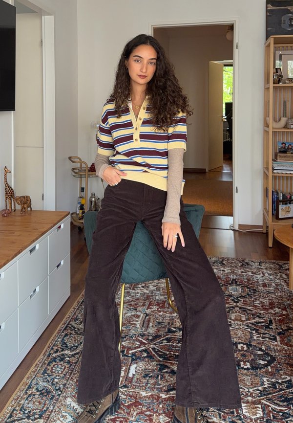 Femme aux cheveux bouclés et foncés portant un haut rayé à manches courtes par-dessus une chemise beige à manches longues et un pantalon large foncé, assise sur une chaise à l'intérieur.