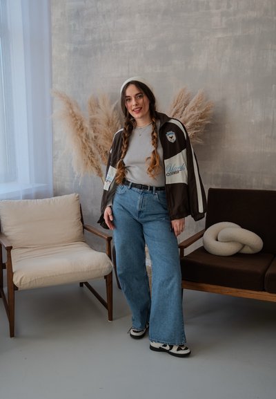 Chaqueta de cierre marrón y azul claro sobre una camiseta gris de canalé, combinada con unos pantalones vaqueros de pierna ancha azules y zapatillas negras. Silla tapizada beige al lado.