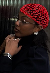 Punainen virkattu huivi, hopeiset ympyrän ja sylinterin muotoiset korut, tumma vaatetus. Lähikuva profiilista, teksturoitu tausta. Silmälasit, joissa on hennot kehykset.