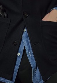 Manteau en laine noire avec deux poches avant et des boutons foncés, porté par-dessus une chemise en jean bleu clair avec des boutons blancs et un accent de couture visible.