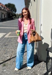 Žena u ružičastom sakou, krem majici i širokim plavim trapericama. Drži zeleni napitak i smeđu kožnu torbicu. Pozadina sa sunčanim ulicama.
