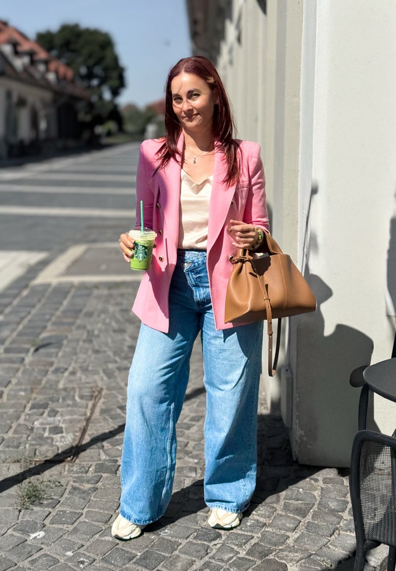 Femme portant un blazer rose, un top crème et un jean bleu à jambes larges. Elle tient une boisson verte et un sac à main en cuir marron. Arrière-plan d'une rue éclairée par le soleil.