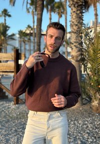 Homme en pull marron et pantalon beige tenant des lunettes de soleil près du visage, debout sur une plage de galets avec des palmiers et une clôture en bois en arrière-plan.