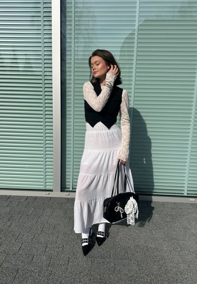 Chaleco negro sobre un vestido largo blanco y de capas. El vestido presenta mangas de encaje floral. Ella lleva un bolso negro con un acento de encaje.