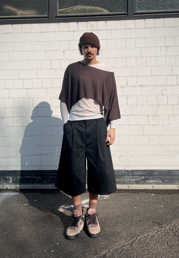 Man wearing brown knit hat, brown poncho over white long-sleeve shirt, wide black cropped pants, and beige sneakers standing against white brick wall.
