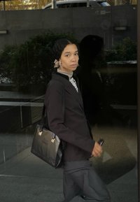 Melnas ādas rokassomi ar zelta aksesuāriem, piegriezta melna žakete, pelēki bikses un gaišas krāsas krekls. Šaurs griezums, gluda tekstūra, minimālas detaļas.