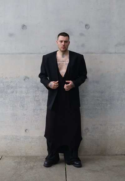 Blazer negro sobre un vestido negro con escote en V profundo. Tatuajes visibles en el pecho y los brazos. Texturas suaves con una paleta de colores oscuros y monocromáticos.