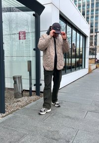 Ανοιχτό μπεζ επενδεδυμένο μπουφάν πάνω από μια μαύρη κουκούλα, ελαιοπράσινα παντελόνια και μαύρα αθλητικά παπούτσια. Το φόντο περιλαμβάνει ένα γυάλινο κτίριο και έναν κάδο απορριμμάτων.