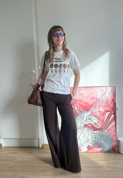 Mujer joven con gafas de sol, camiseta blanca con diseño gráfico, pantalones a rayas, lleva un bolso marrón, de pie junto a una pintura de unicornio roja y gris en interiores.