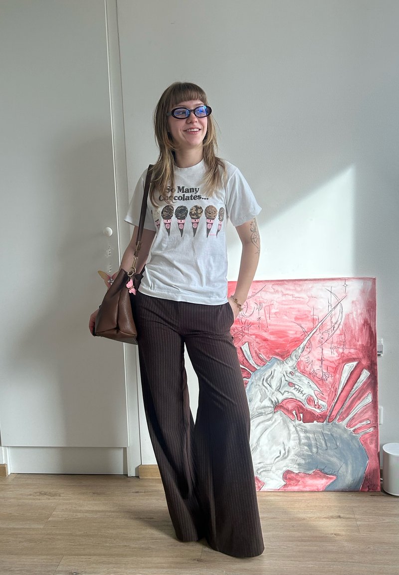 Tânără purtând ochelari de soare, tricou alb cu grafică, pantaloni cu dungi, ținând o geantă maro, stând lângă o pictură cu un unicorn roșu și gri în interior.