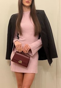 Femme portant une robe côtelée rose aux manches évasées, un blazer noir posé sur les épaules, tenant un sac à main bordeaux avec une fermeture dorée.
