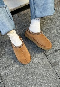 Bēša zamšādas slip-on stila apavi ar rakstainu adījuma apdari apkārt apkaklei, gumijas zolīte un apaļas formas purngalu, valkāti ar baltām zeķēm.
