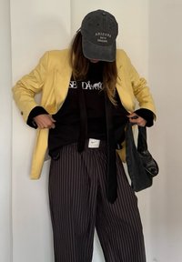 Personne portant un blazer jaune, une casquette noire, un pull graphique noir, un pantalon rayé et tenant un sac à main noir devant un fond uni.